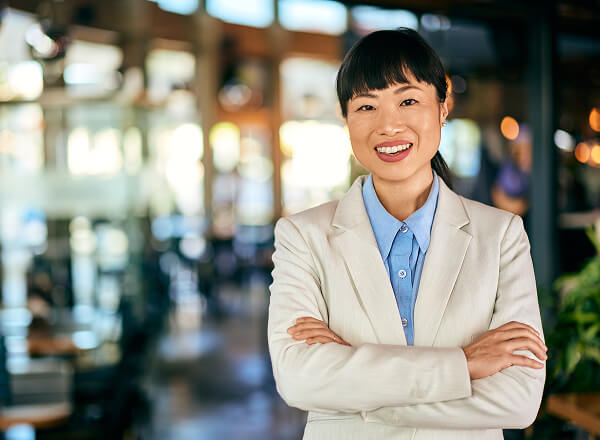 A woman in a light-colored suit with arms crossed smiles at the camera in a modern indoor setting.