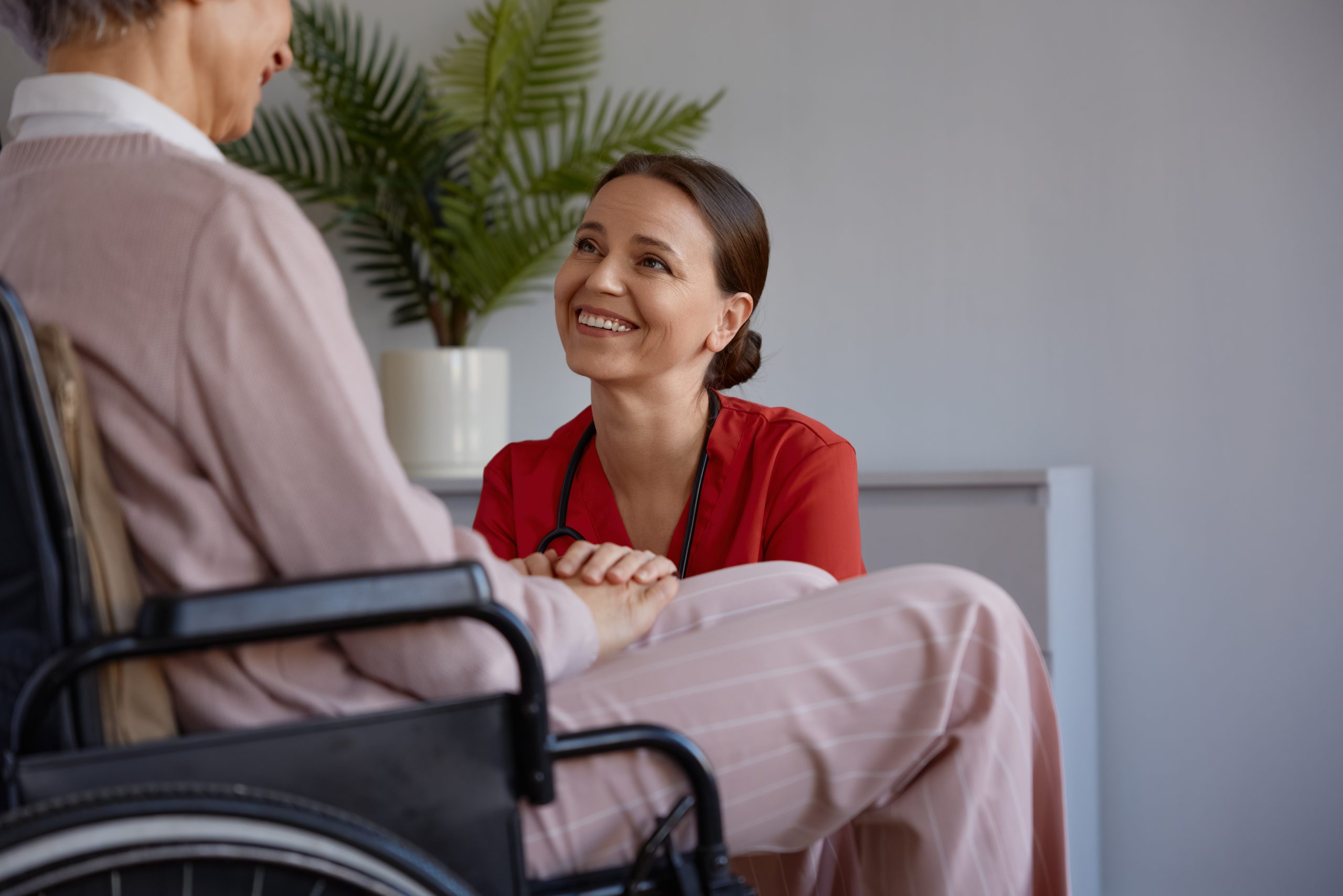 A woman in a red shirt smiles and holds hands with an older person sitting in a wheelchair indoors, with a potted plant in the background.